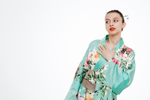 Woman in traditional japanese kimono looking aside holding hand on her chest feeling thankful on white