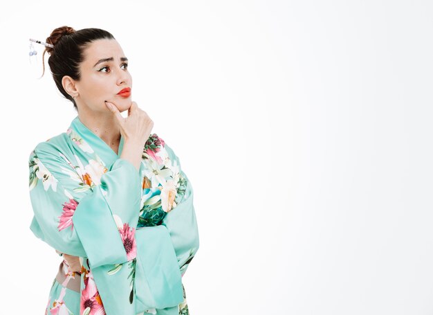 Woman in traditional japanese kimono looking aside with pensive expression thinking holding hand on her chin on white