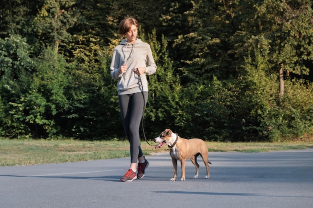 Woman trains her dog to run along during jogging. Young fit female and staffordshire terrier dog at a morning walk doing physical excercise