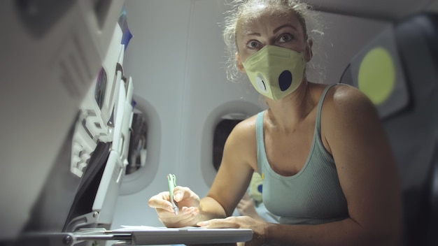 Woman travel caucasian at plane with wearing protective medical mask Girl tourist at aircraft with protect respirator fill out a registration form about her health Coronavirus sarscov2 covid19