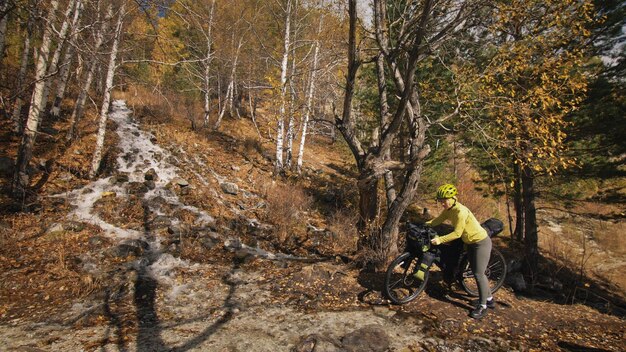 女性はバイクパッキングで混合地形サイクルを旅行します自転車バッグを持った旅行者の旅緑の黒い色のスポーツウェア魔法の秋の森の川の小川での旅行