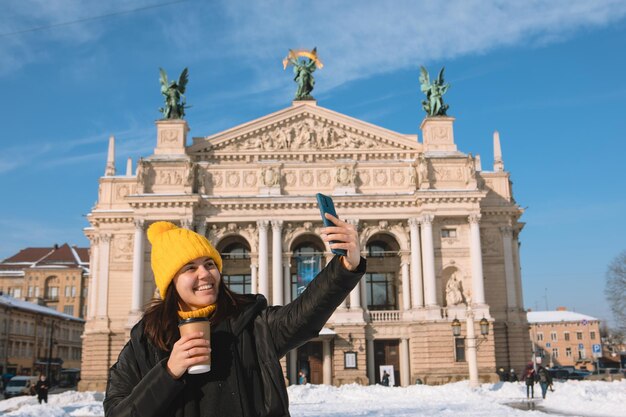 Woman traveler drinking coffee to go taking selfie in front of opera building lviv city ukraine