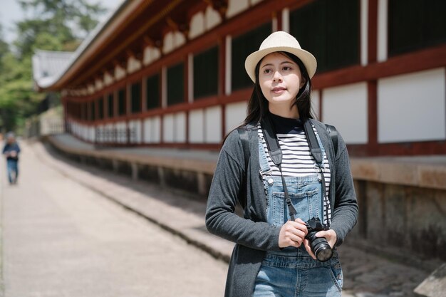 높은 구조의 일본 전통 건물 근처를 바라보는 여성 여행자. 카메라를 들고 있는 아시아 여성은 화창한 날 멀리 아름다운 경치를 감상하고 있습니다.