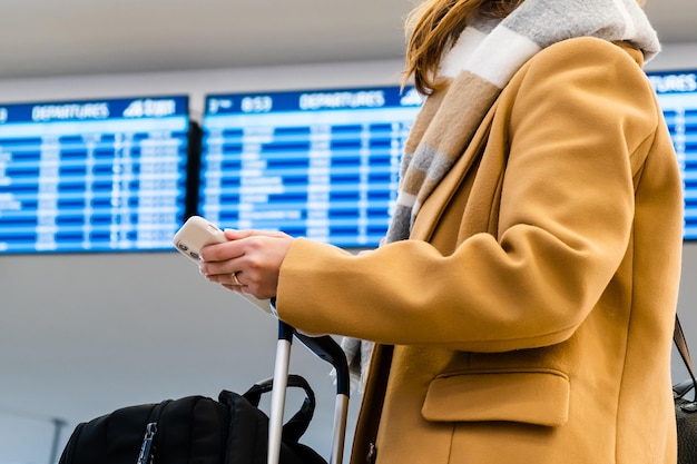 暖かいスカーフとスタイリッシュな黄色のコートを着た女性旅行者は、携帯電話とスーツケースを再び立っています...
