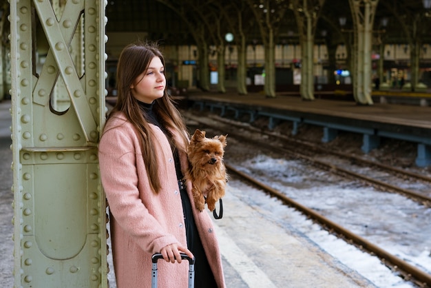 여성 여행자 관광객은 분홍색 코트를 입고 기차역에서 수하물과 개를 들고 산책합니다.