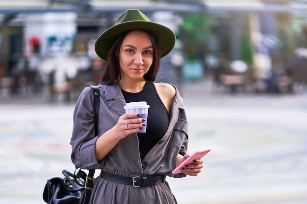 Woman traveler wearing felt hat and overalls with a backpack holds paper cup