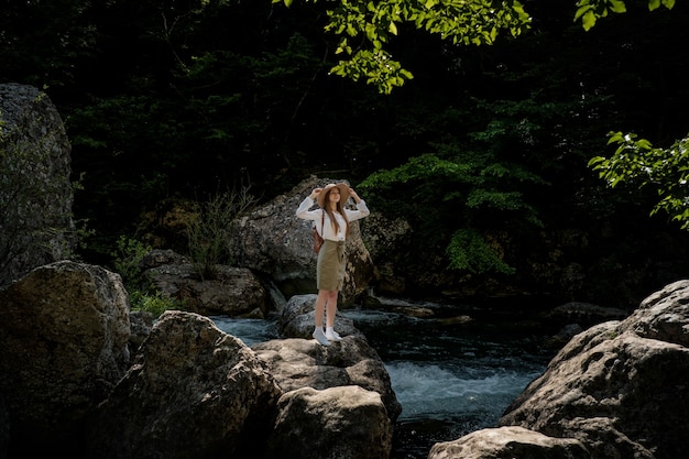 배낭과 모자 깊고 푸른 물과 함께 강 근처 놀라운 산과 숲에서 산책하는 여자 여행자
