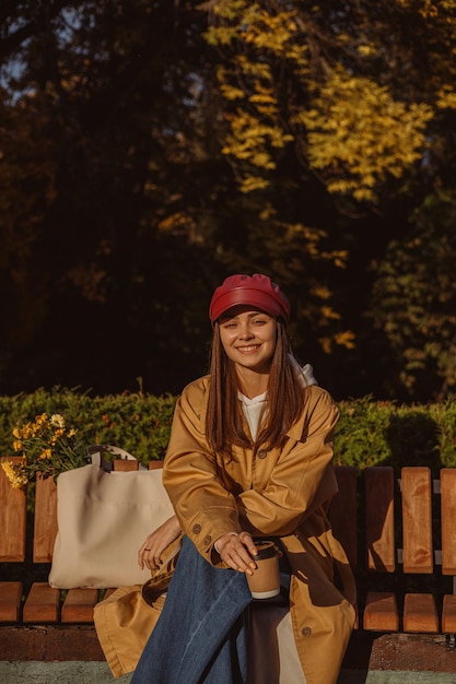 woman in trendy trench and cap with cup of takeout coffee relaxing on bench on sunny day