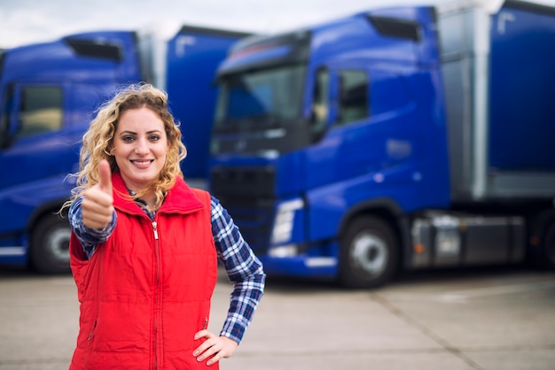 Woman truck driver in casual clothes holding thumbs up in front of truck vehicles.