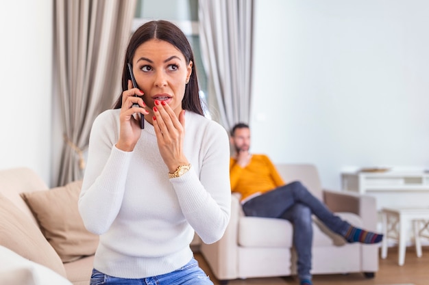 Woman turned her back to man, talking on phone with her lover, boyfriend sitting in the back watching tv.