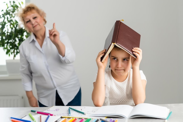 Woman tutor or foster parent mum helping cute caucasian school child girl doing homework sitting at table. Diverse nanny and kid learning writing in notebook studying at home.