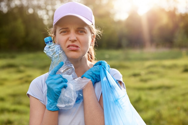 Woman twisted her face, holding used plastic bottle and garbage bag, being tired of cleaning up parks, calls on to reuse waste, do not pollute environment.