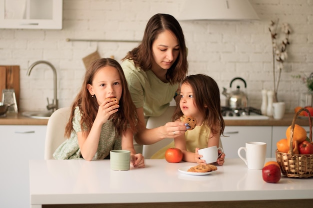 A woman and two young girls sit at a kitchen table eating cookies