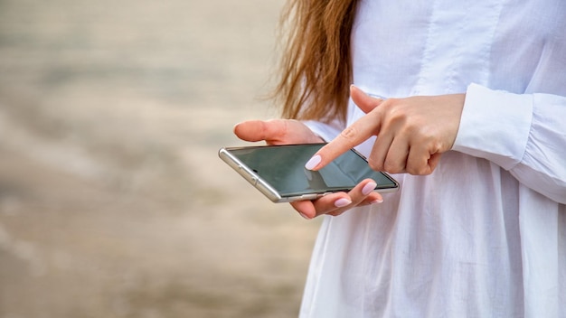 La donna usa il telefono cellulare per cercare in internet o controllare l'app all'esterno. mani femminili del primo piano con il resto dello smartphone al fondo dell'acqua di mare. messaggio di testo toman in viaggio. banner web lungo con spazio di copia.