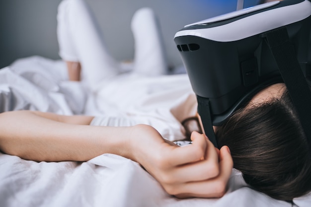 여자는 침대에서 편안하게 집에서 가상 현실 안경을 사용합니다. 미래의 게임, 기술 플레이