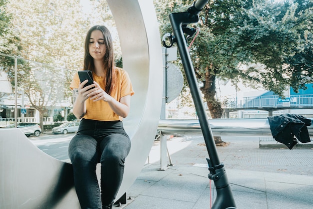 Woman uses her smartphone while moving with her electric scooter around the city street New ecofriendly transport woman using phone cell for unlocking electric push scooter