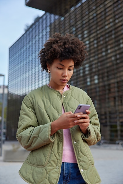 Photo woman uses mobile phone for sending messages searches web publication connected to wireless internet dressed casually poses in city centre uses chat application