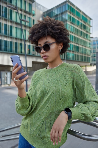 女性はスマートフォンを使用してニュースをオンラインで読むサングラスをかけている緑のジャンパーはワイヤレスインターネット接続を介してメールを送信します