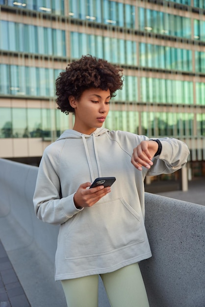 woman uses smartwatch connected via bluetooth to phone checks her activity after sport training wears sweatshirt and leggings poses against modern glass city architecture