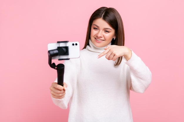 Woman using cell phone and steadicam for broadcasting livestream pointing down asking to subscribe wearing white casual style sweater Indoor studio shot isolated on pink background