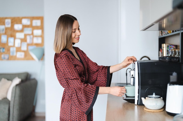 Woman using coffee machine to make big mug of coffee at home Woman wearing silk robe at home while preparing a latte