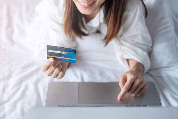 Woman using computer laptop and credit card for online shopping while making order on bed in morning at home technology ecommerce digital banking online payment and apartment living concept