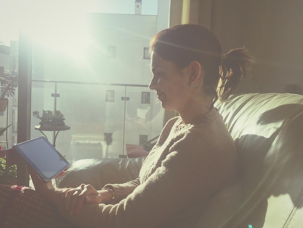 Woman using digital tablet while sitting on sofa at home during sunny day