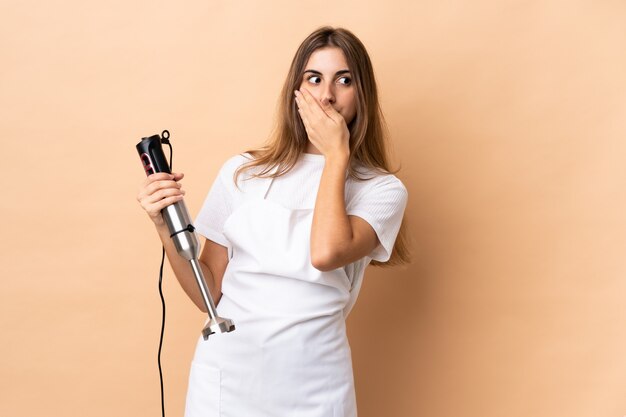 Woman using hand blender over isolated wall doing surprise gesture while looking to the side