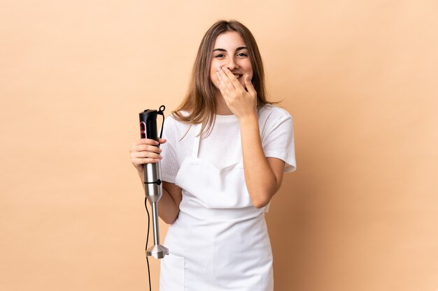 Woman using hand blender over isolated wall happy and smiling covering mouth with hands