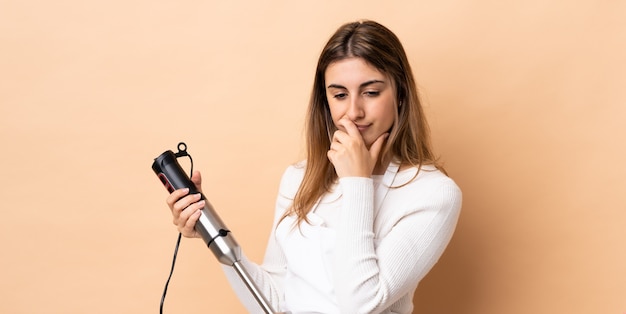 Woman using hand blender over isolated wall having doubts