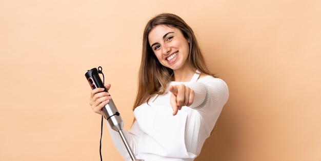 Woman using hand blender over isolated wall pointing front with happy expression