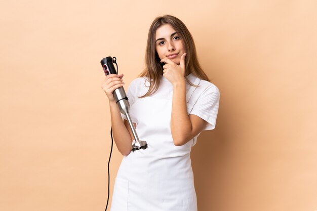 Woman using hand blender over isolated wall thinking