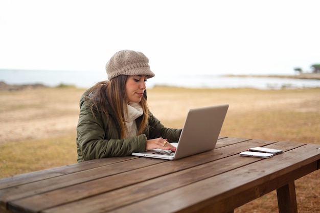 Woman using internet connection outdoor, sitting at a wooden table in a sea park working at laptop