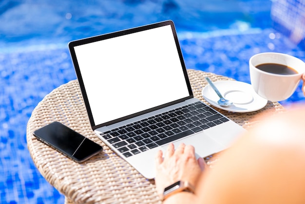 プールの空の白い画面のモックアップでコーヒーを飲みながら、休暇中にラップトップ コンピューターを使用している女性