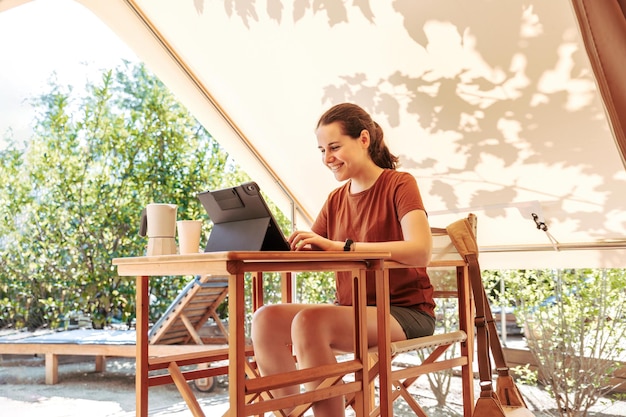 Женщина, использующая ноутбук в уютной палатке для кемпинга в солнечный день, роскошная палатка для кемпинга для летнего отдыха