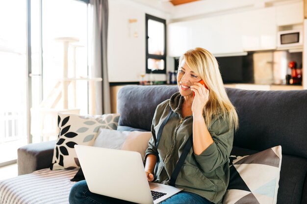 Donna che usa un laptop e conversa su uno smartphone rosso su un divano