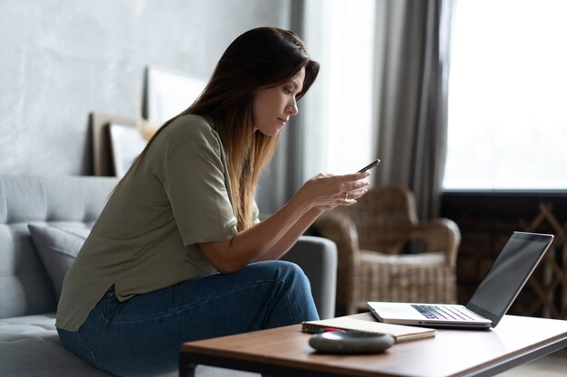 ノートパソコンと携帯電話をソファで使用している女性。