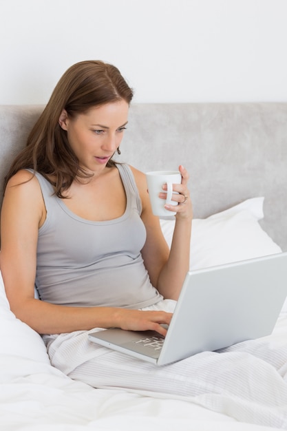 Donna che per mezzo del computer portatile mentre mangiando caffè a letto