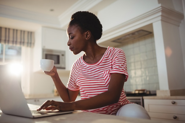 キッチンでコーヒーを飲みながらノートパソコンを使用している女性