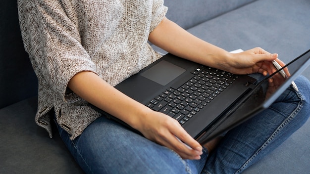 Женщина, использующая ноутбук с пустым экраном, сидя на диване в домашнем интерьере, вид сзади