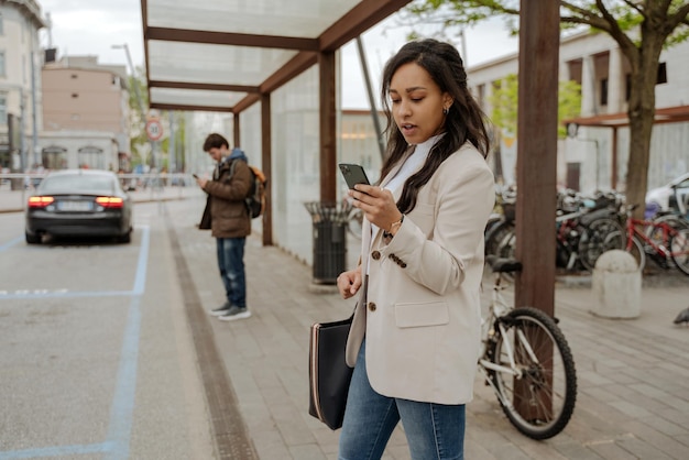 Фото Женщина, пользующаяся мобильным телефоном онлайн, читающая текстовое сообщение, стоящее на автобусной остановке