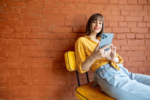 Женщина с помощью смартфона, сидя на стуле на фоне кирпичной стены