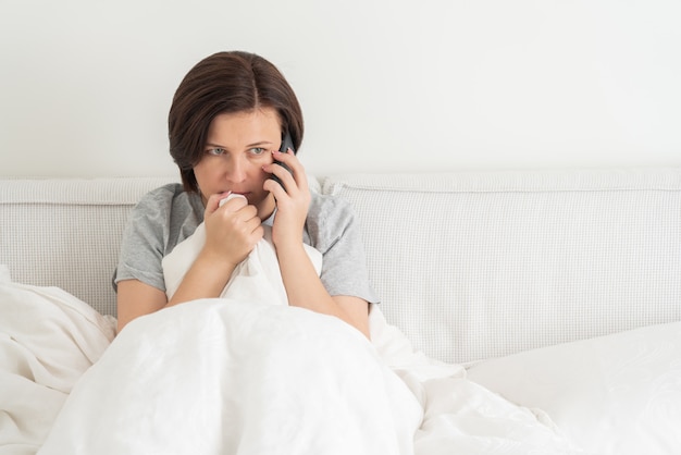 Woman using smartphone as she sits in bed covered with duvet, scared and terrified