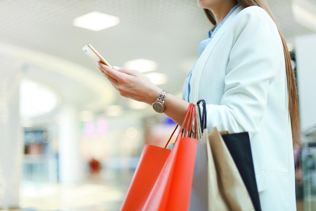 스마트폰을 사용하고 쇼핑몰 배경에 서 있는 동안 쇼핑백을 들고 있는 여자.