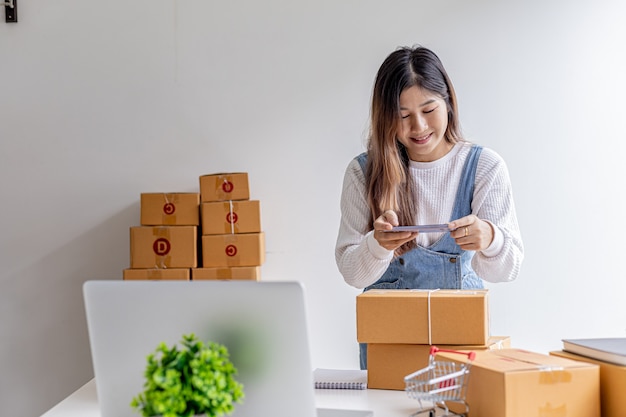 A woman using a smartphone to take pictures in front of parcel boxes, parcel boxes for packing goods, delivering goods through private courier companies. Online selling and online shopping concepts.