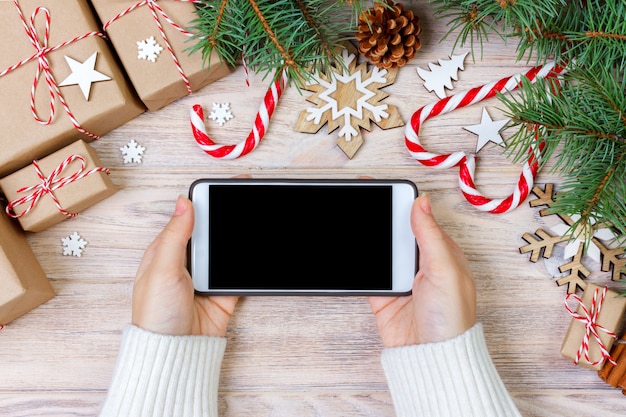 Donna che per mezzo dello smartphone con lo schermo in bianco, la struttura festiva del trumpery, la ricerca del regalo di natale, lo shopping online, gli sconti stagionali e il concetto di vendita