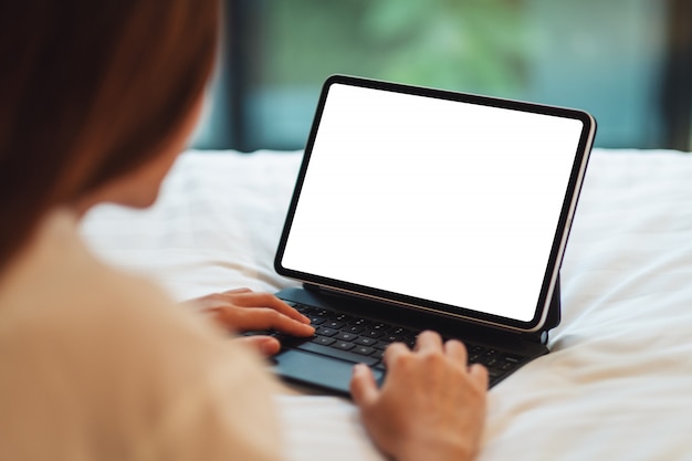  of a woman using and typing on tablet pc with blank white desktop screen as computer pc while lying down on a cozy white bed at home