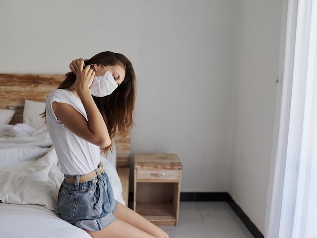 Woman on vacation with medical mask on her face sits on bed in isolated room