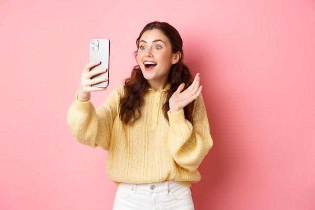 Woman video chatting with surprised face, receive amazing news, gasping excited, seeing something in smartphone screen, standing against pink wall.