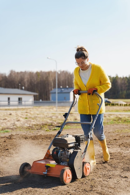 Woman villager is using aerator machine to scarification and aeration of lawn or meadow